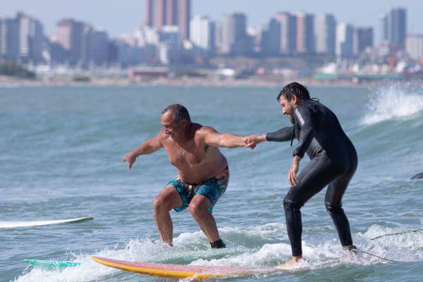 Iniciante surfista executando um take off em uma onda suave, com orientação de um instrutor ao fundo. A imagem é a capa do artigo Manobras Básicas de Surf: Guia para Iniciantes.