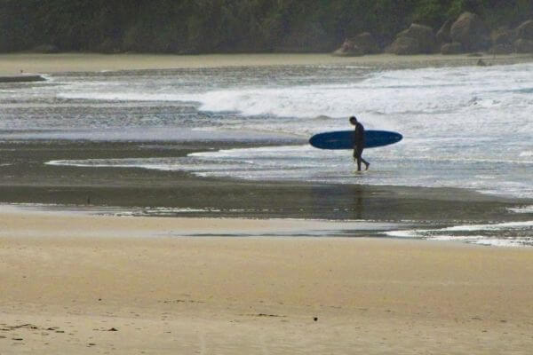 Um homem saindo do mar com sua prancha debaixo do braço. a imagem é a capa do artigo, as Melhores Praias para Surfar no Brasil: De Ubatuba a Fernando de Noronha