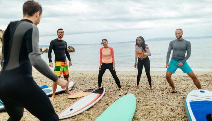 Instrutor de surf na praia com 4 alunos, 2 mulheres e 2 homens, com o mar ao fundo, preparando-se para uma sessão de treino. Imagem ilustra o artigo 5 Melhores Exercícios de Treino Funcional para Surfistas: Melhore Sua Performance