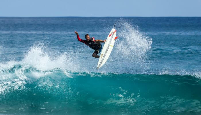 Surfista profissional executando um aéreo 'air reverse' com uma grande onda azul ao fundo.