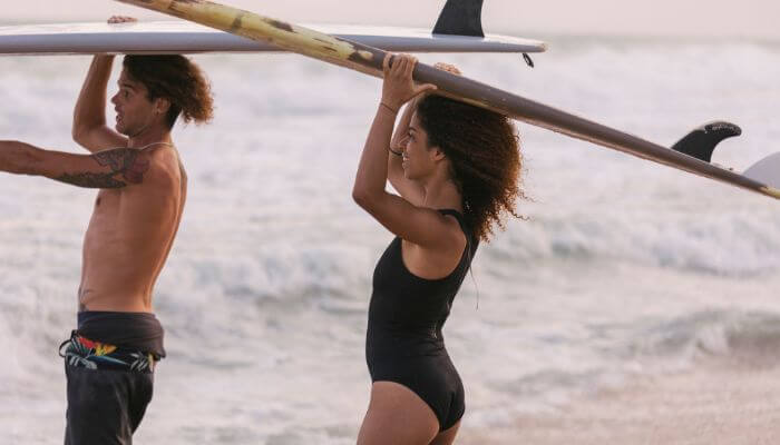 Dois surfistas, um homem e uma mulher, com pranchas na cabeça na beira da praia, homem aponta para uma onda.